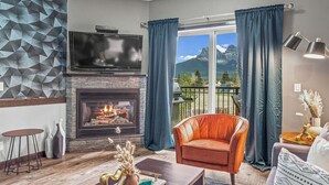 A living room with a fireplace, a television, and a view of mountains through large windows. - Experience Cozy Rockies Escape w/ Pool & Hot tub (Canmore)
