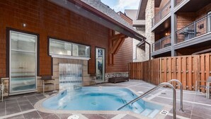 A hot tub area with a stone wall, wooden deck, and a balcony with railings. - Experience Cozy Rockies Escape w/ Pool & Hot tub (Canmore)