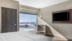 Una habitación de hotel moderna con una cama grande, un televisor en la pared, una silla y vistas de la ciudad a través de un ventanal amplio.
