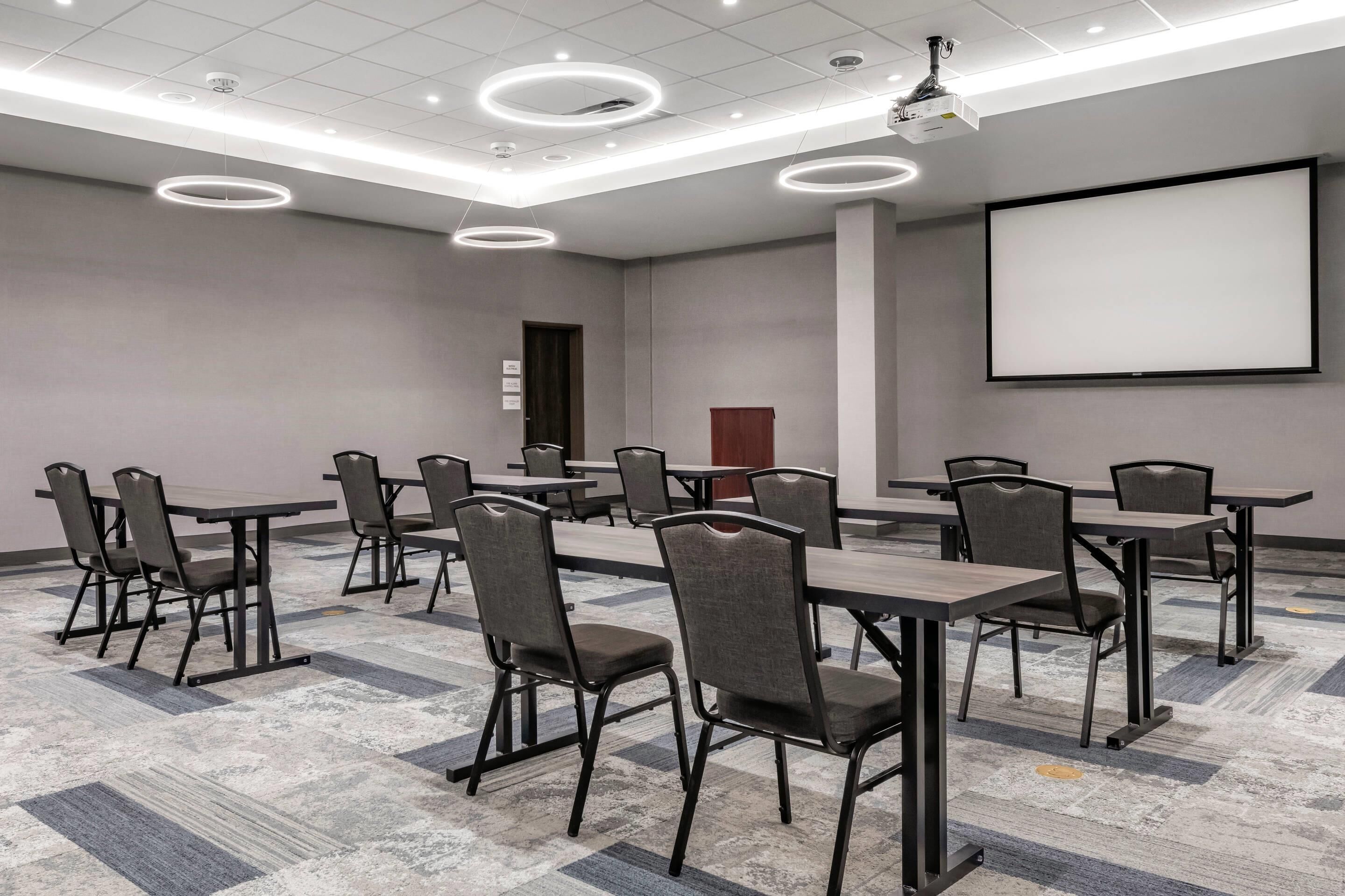 A conference room with tables and chairs, a projector screen, and a carpeted floor.