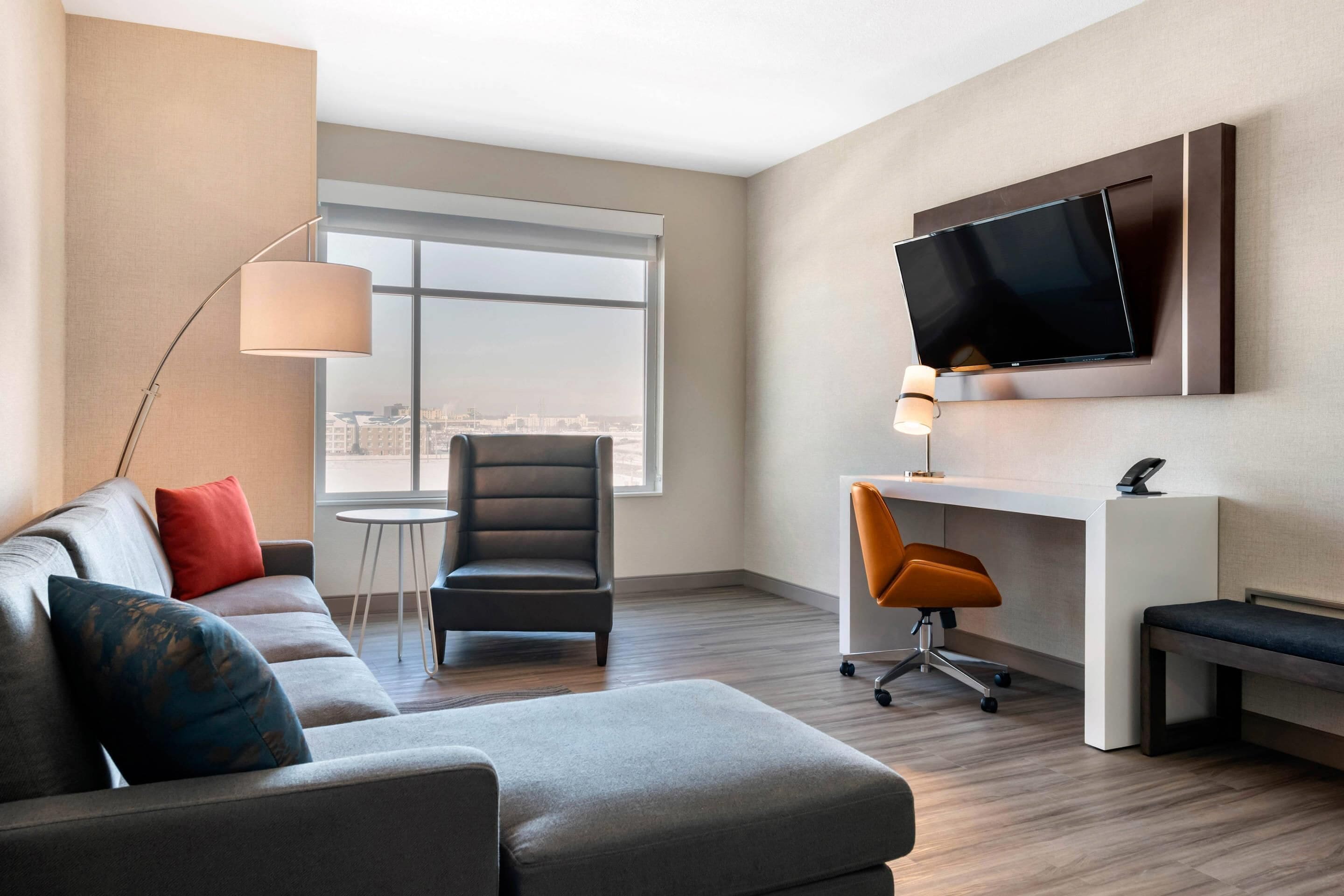 Una habitación de hotel moderna con sofá, escritorio, silla, televisor montado en la pared y un gran ventanal con vistas a la ciudad.