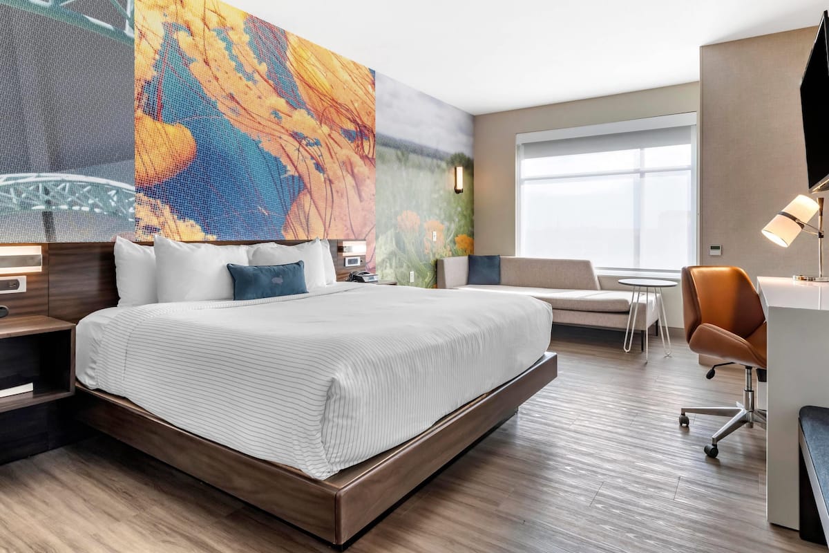 A modern hotel room with a large bed, a desk, a chair, and a large window.