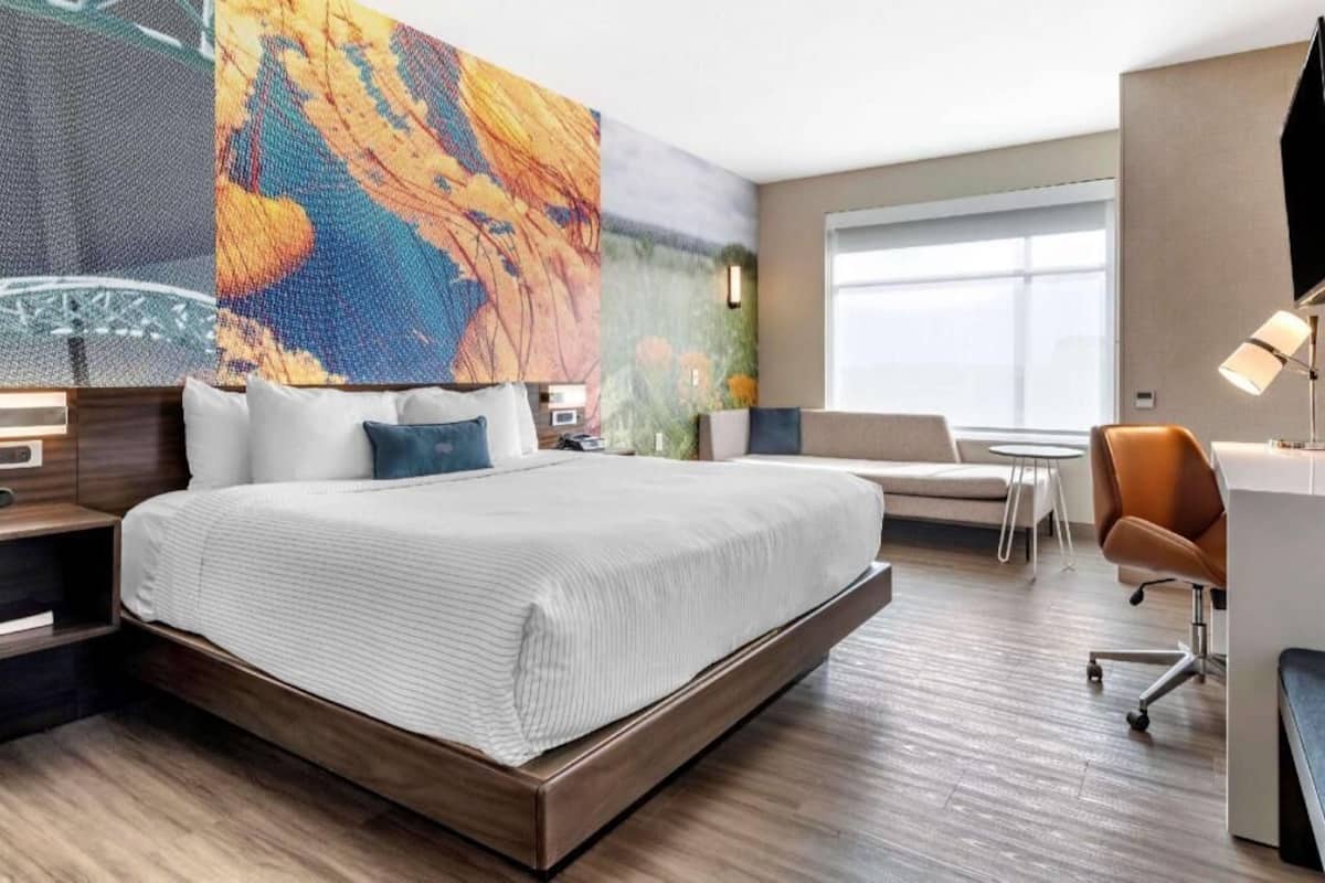 A modern hotel room with a large bed, a desk, a chair, and a large window.