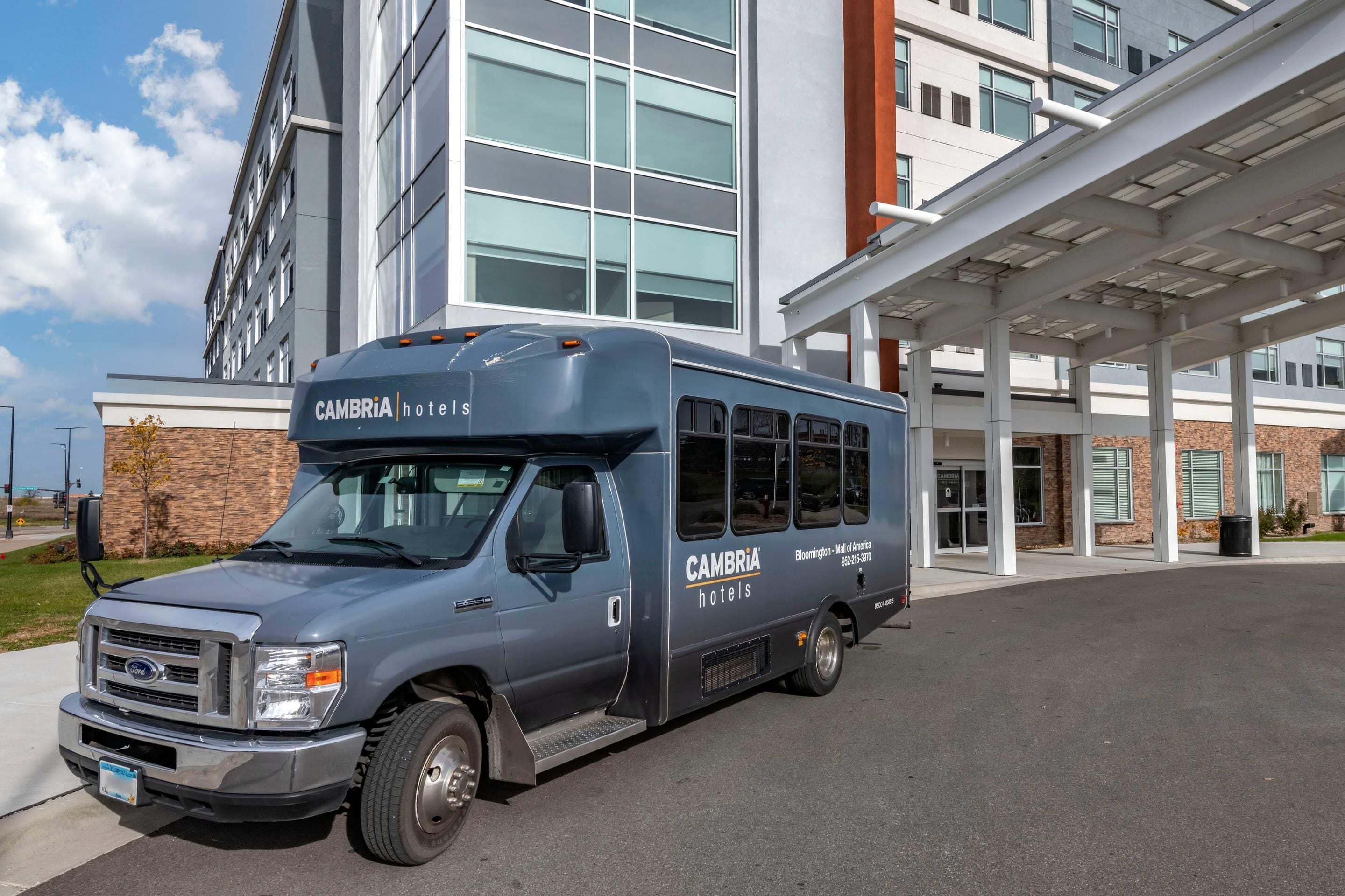 Un autobús azul con la marca 'CAMBRIA hotels' estacionado frente a la entrada de un hotel.
