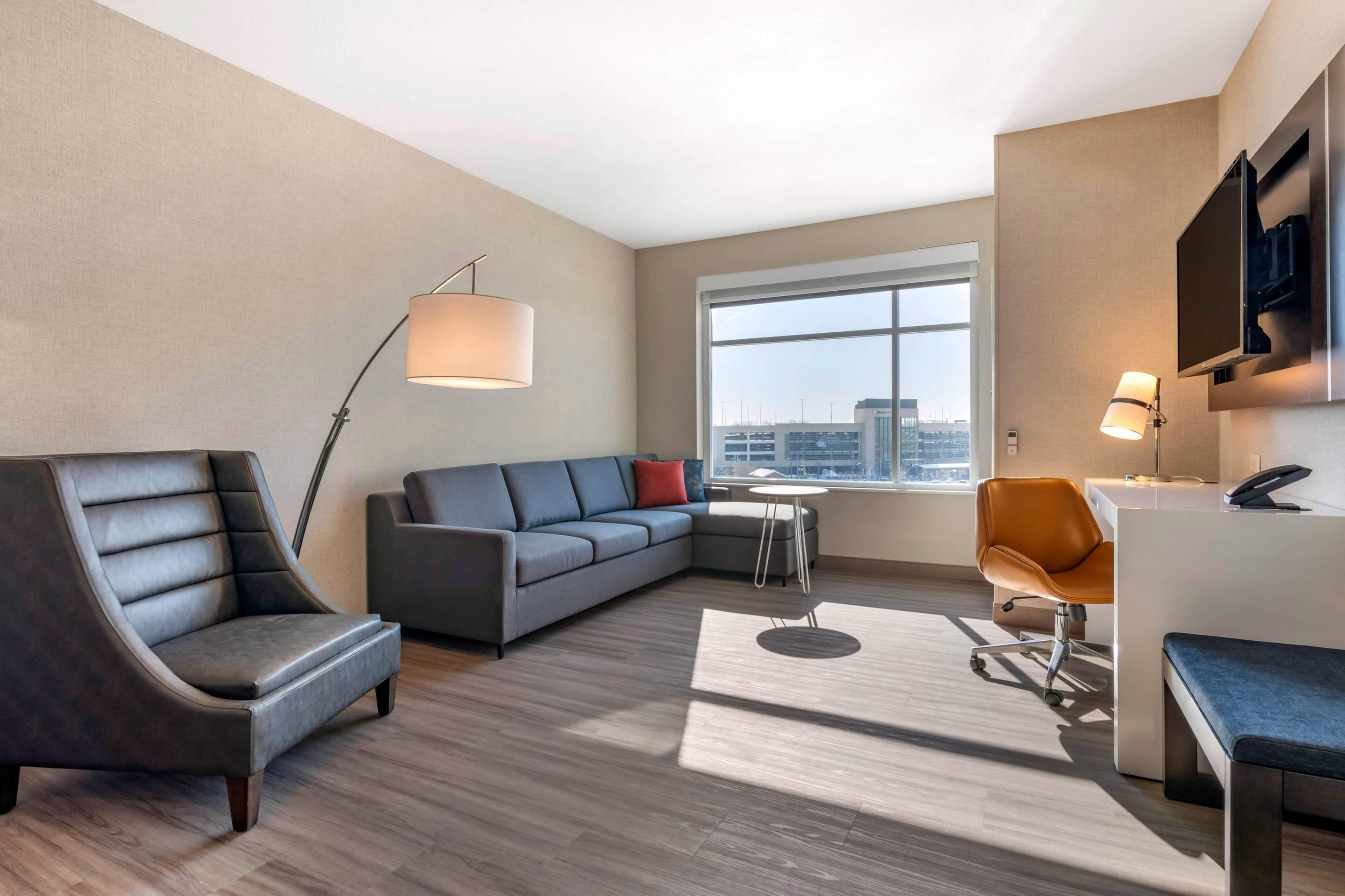 Une chambre d’hôtel moderne équipée d’un canapé, d’un fauteuil, d’un bureau et d’une grande fenêtre donnant sur la ville.