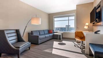 A modern hotel room with a sofa, armchair, desk, and a large window offering a city view.
