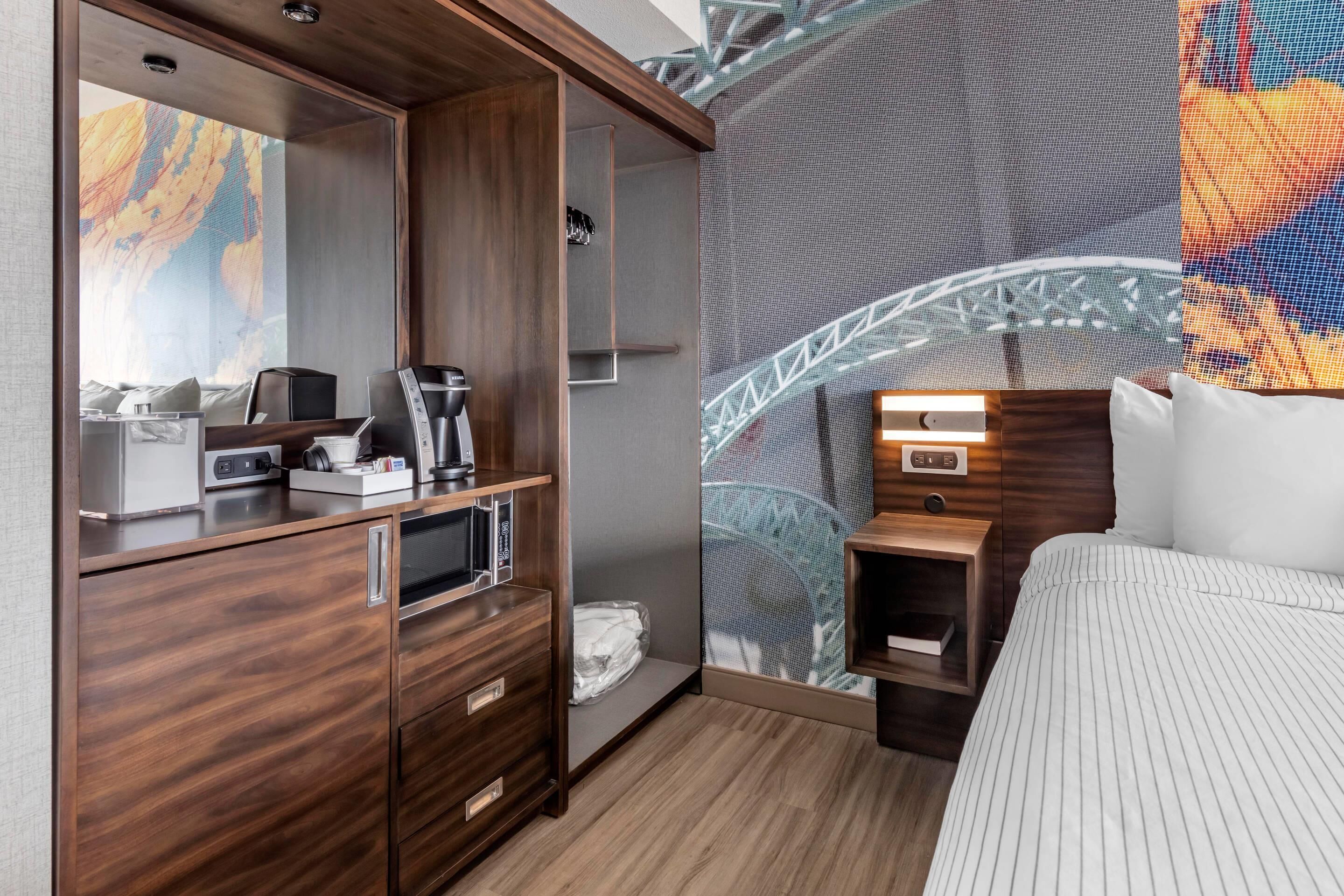 A hotel room with a wooden wardrobe, a bed with striped bedding, a small bedside table, a built-in microwave, and a large window with a view of a bridge.