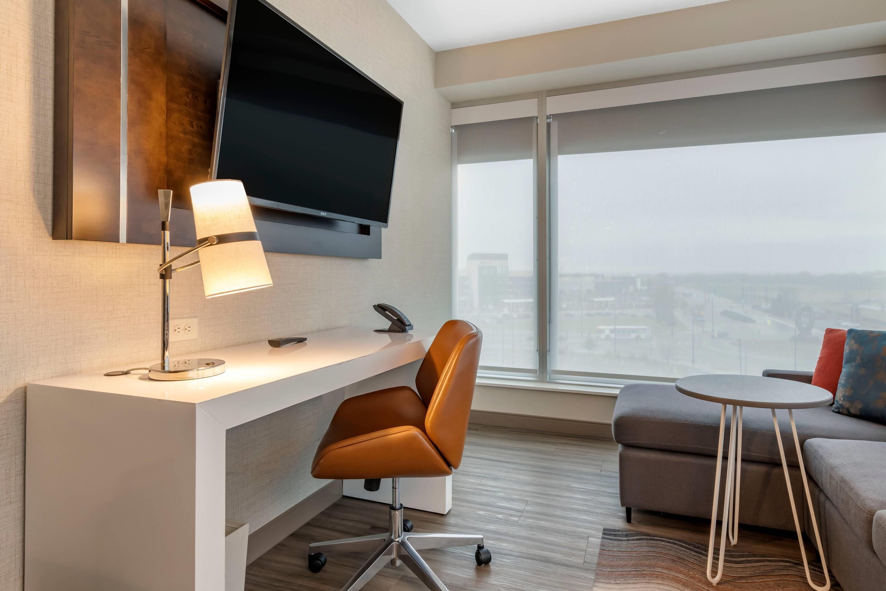 A modern hotel room with a desk, chair, television, and a large window with a city view.