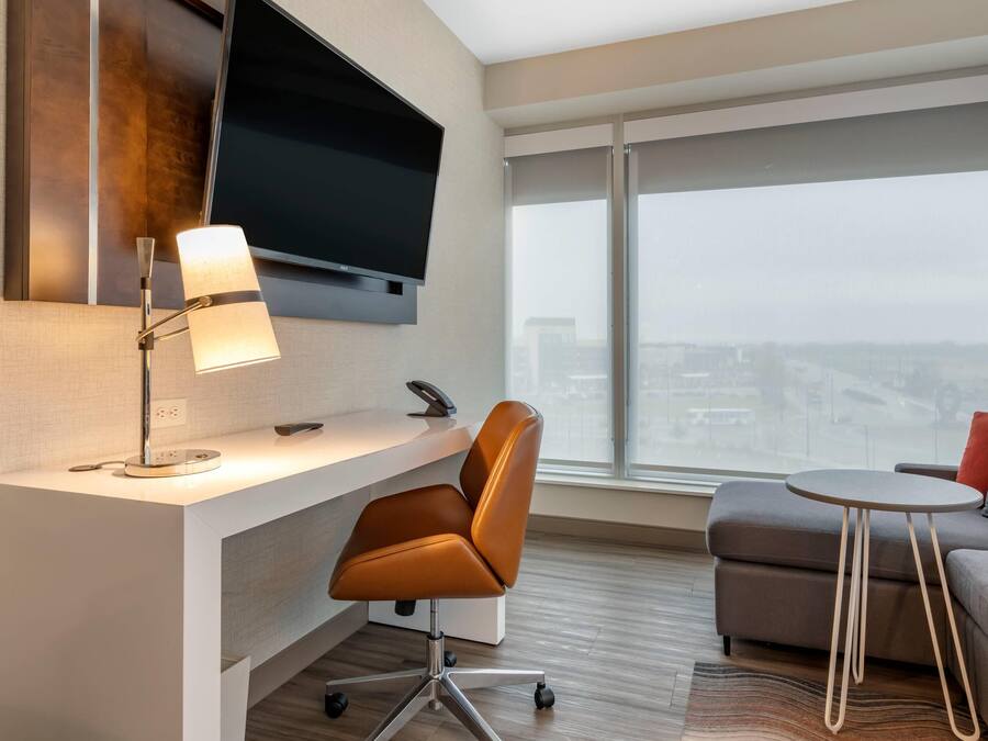 A modern hotel room with a desk, chair, television, and a large window with a city view.