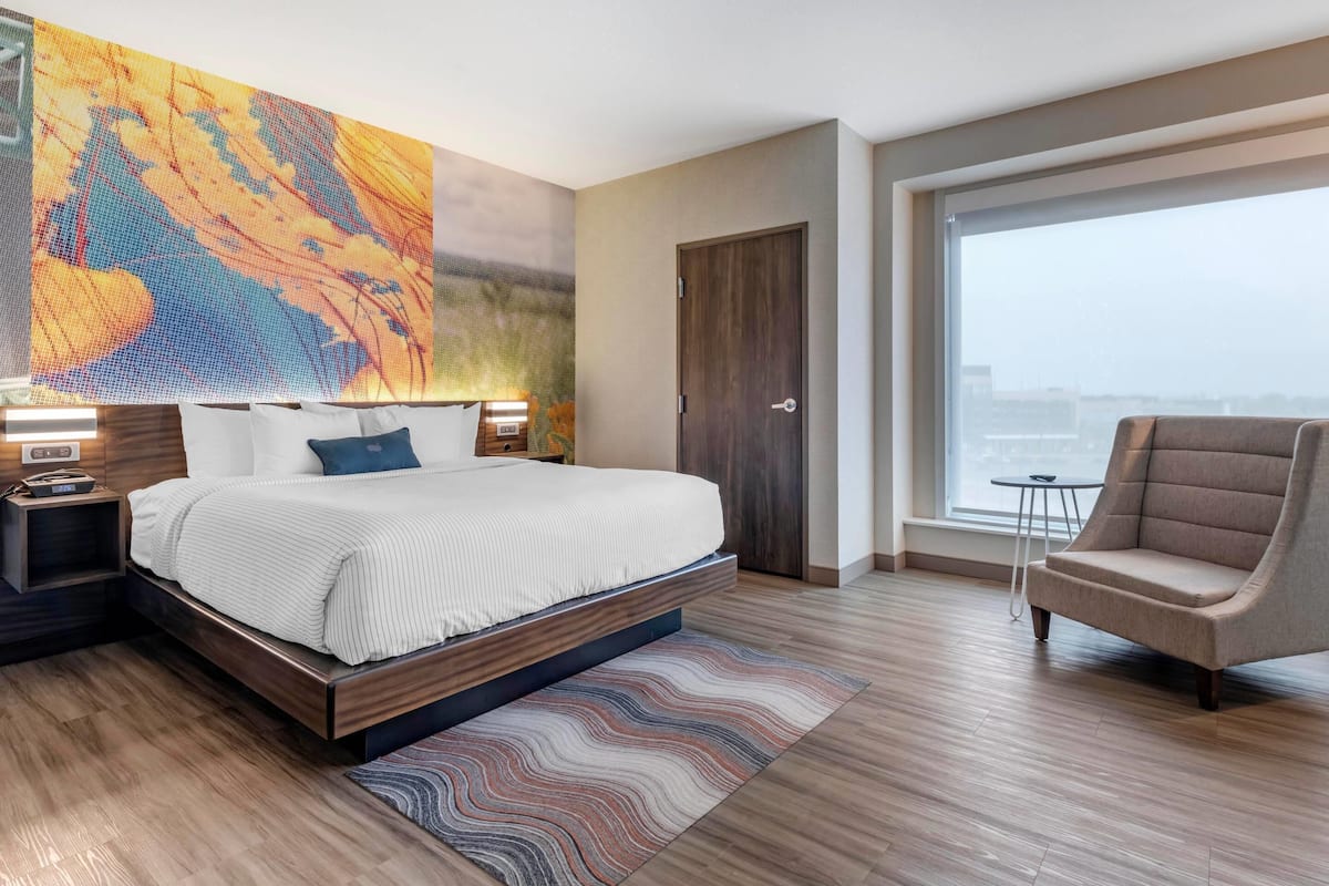 A modern hotel room with a large bed, a chair, a view through a large window, and a wall art piece.