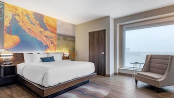 A modern hotel room with a large bed, a chair, a view through a large window, and a wall art piece.