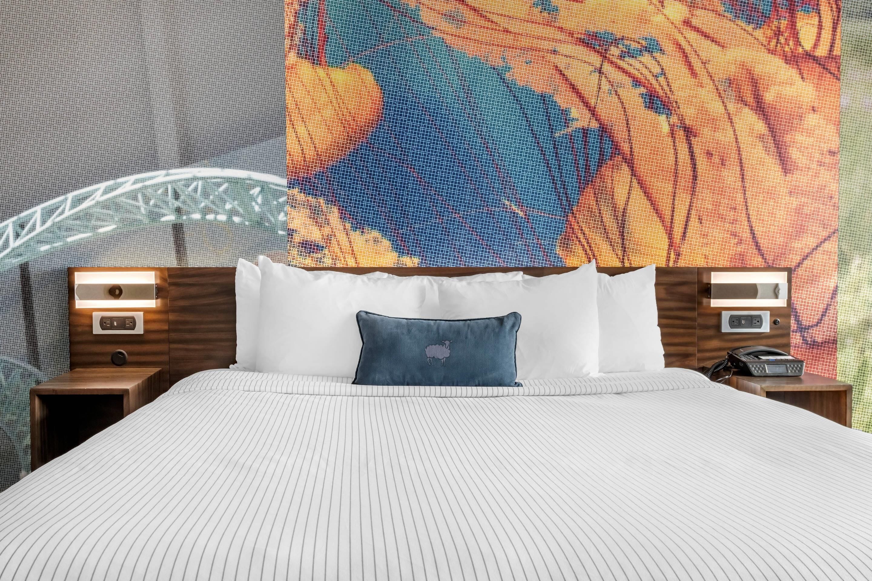 Una cama bien hecha con edredón blanco a rayas, un cojín azul con diseño de oveja y dos mesitas de noche con lámparas y un teléfono.
