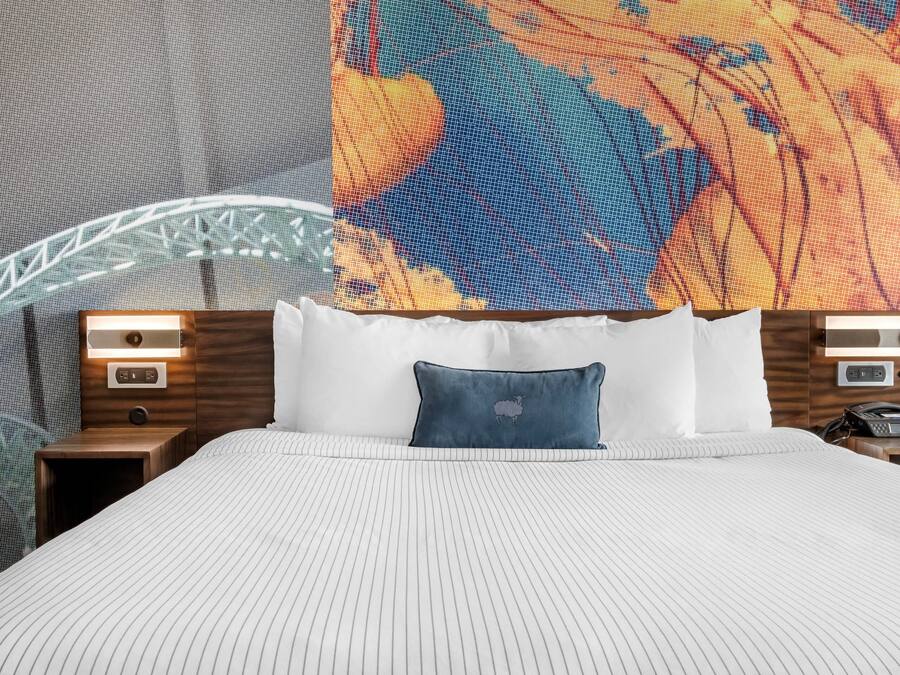 Una cama bien hecha con edredón blanco a rayas, un cojín azul con diseño de oveja y dos mesitas de noche con lámparas y un teléfono.