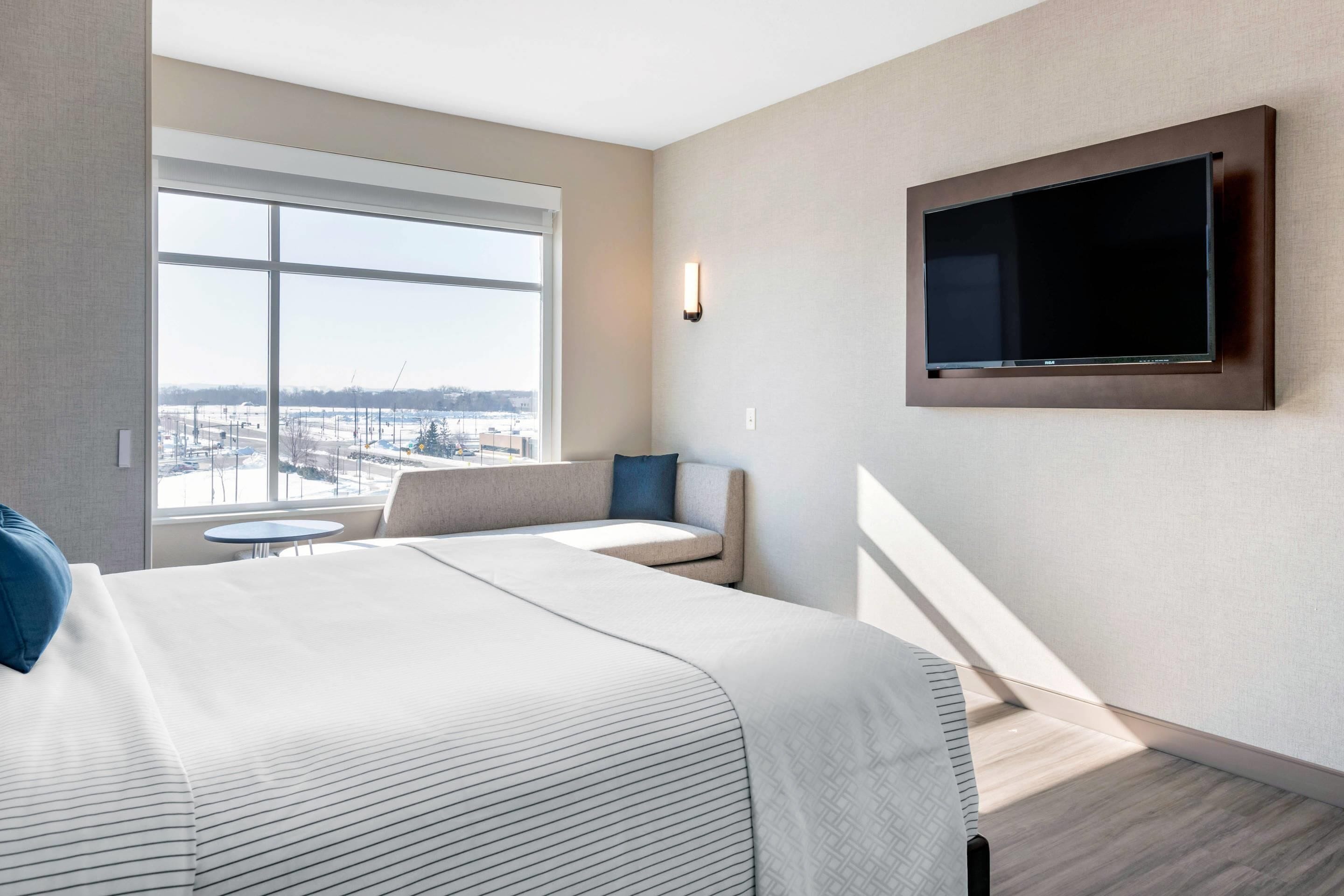 Ein Hotelzimmer mit einem großen Bett, einem an der Wand befestigten Fernseher, einem kleinen Tisch und Blick auf die Stadt durch das Fenster.