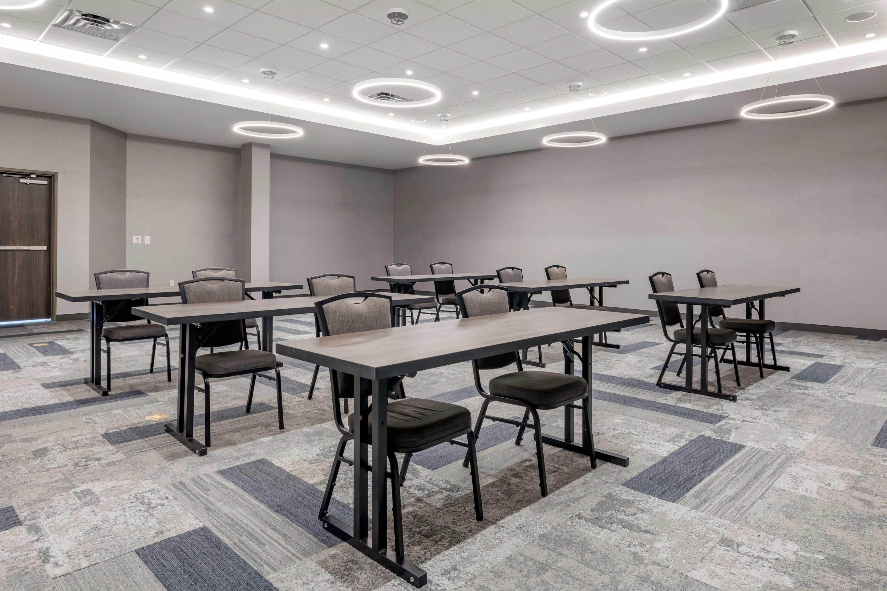 Une salle de conférence dotée de plusieurs tables et chaises, d’un tapis à motifs et de luminaires circulaires au plafond.