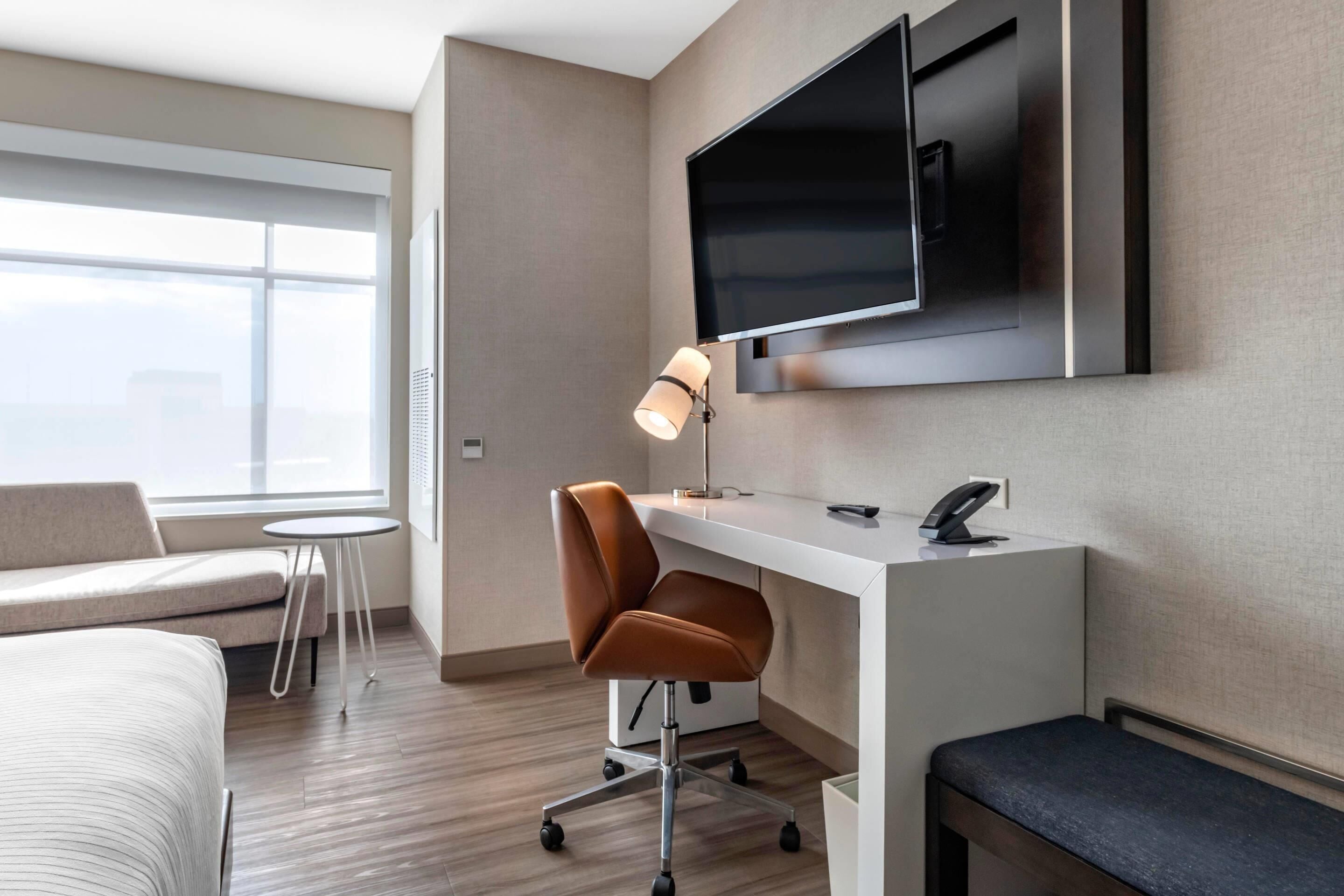 A hotel room with a bed, a desk, a chair, a TV mounted on the wall, a lamp, and a window with a view of the city.