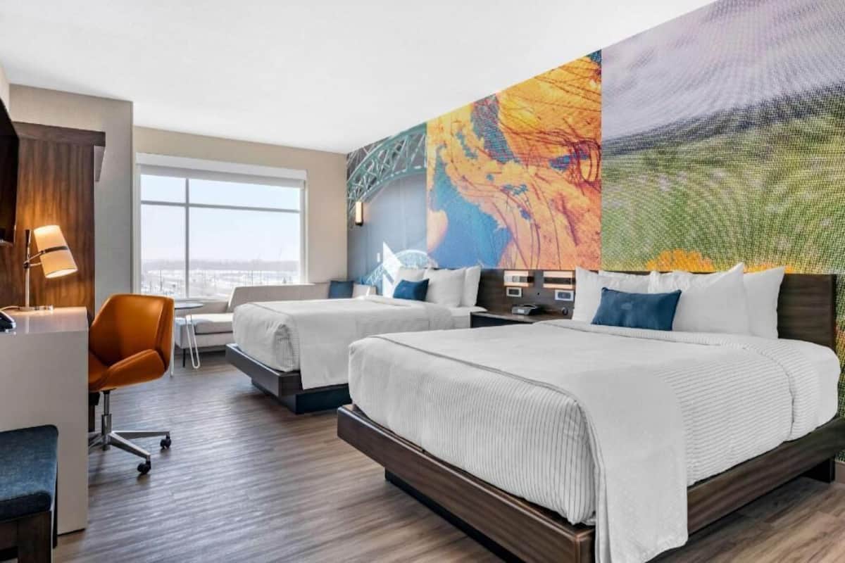 A hotel room with two beds, a desk, a chair, and a large window.