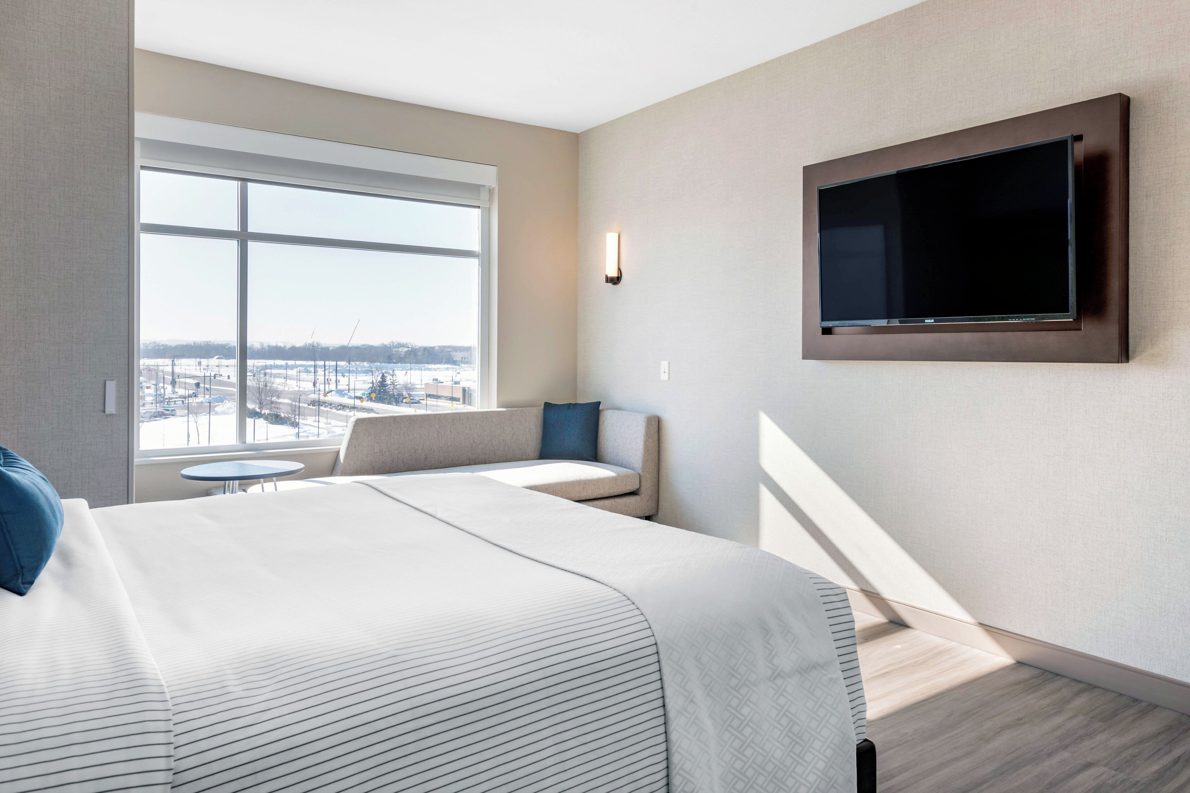 A modern hotel room with a large bed, a seating area with a small table, a TV mounted on the wall, and a view of the cityscape through a large window.