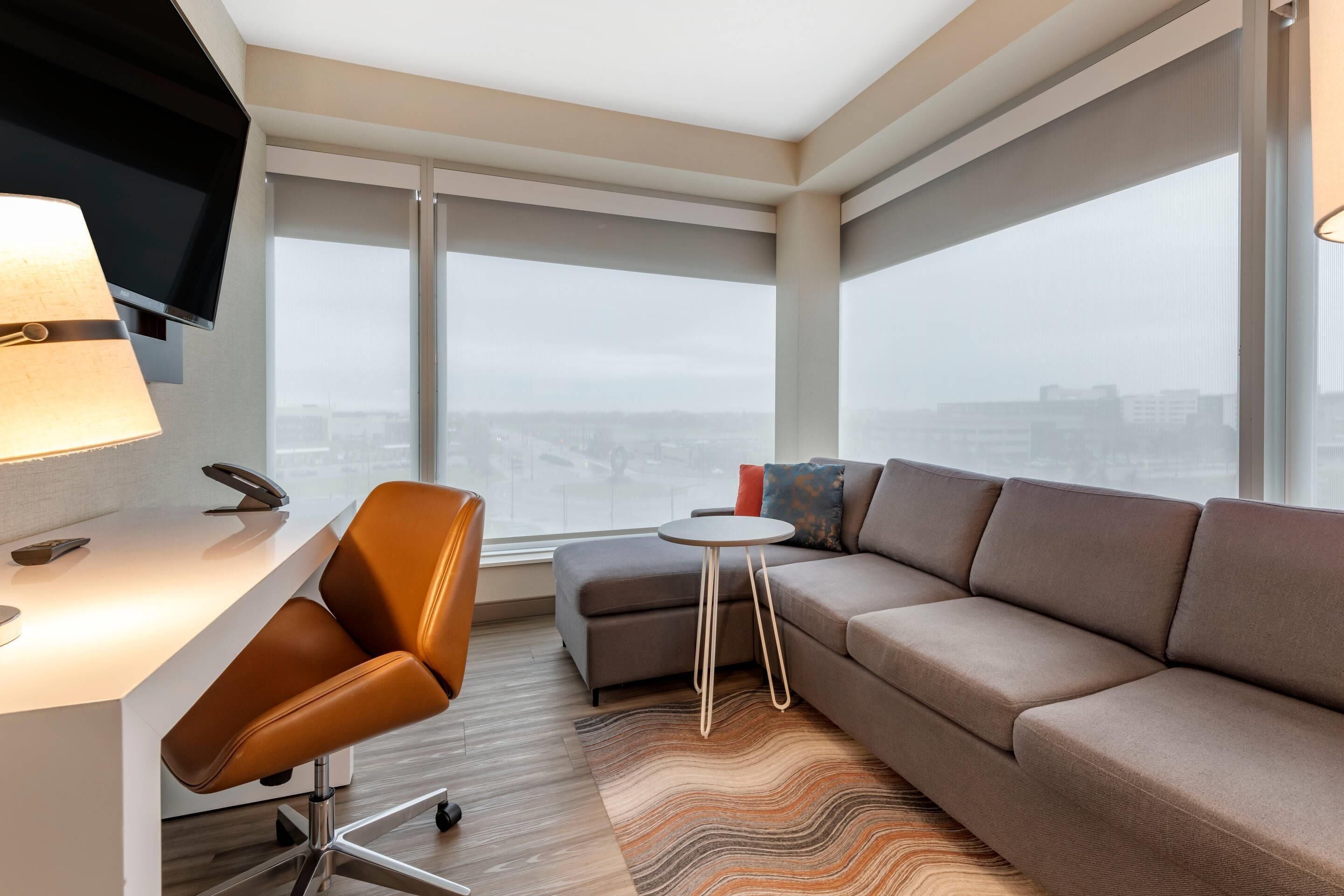 Una habitación de hotel moderna con un ventanal grande, un sofá, una mesita y un escritorio con lámpara.