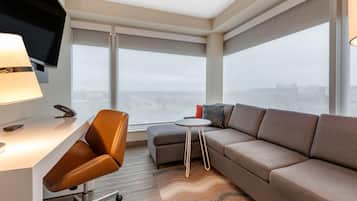 Quarto de hotel moderno com uma janela grande, um sofá, uma mesa pequena e uma escrivaninha com abajur.