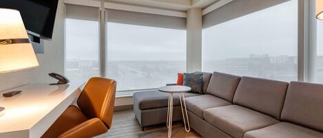 Una habitación de hotel moderna con un ventanal grande, un sofá, una mesita y un escritorio con lámpara.