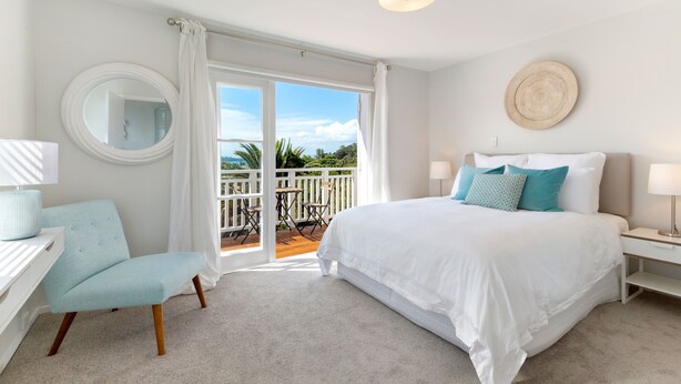 A bedroom with a large bed, a chair, a desk, a mirror, and a balcony with a view of greenery. - Eight on Church Waiheke Accommodation (Waiheke Island)