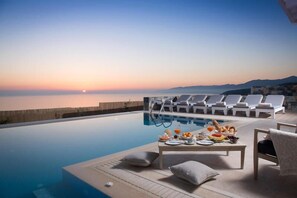Ein Poolbereich mit Liegestühlen und einem Tisch, gedeckt mit Frühstücksangeboten, mit Blick auf ein Gewässer bei Sonnenuntergang.