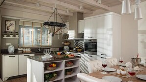 A modern kitchen with white cabinetry, a central island with baskets of fruits and vegetables, and a dining area with a table set for two. - Villas Crete (Hersonissos)