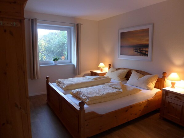 Un lit en bois avec deux oreillers et une couverture, une fenêtre avec des rideaux, une table de chevet avec une lampe et un tableau encadré au mur.