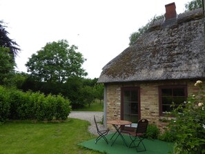 Une petite maison au toit de chaume, aux murs en briques, dotée d’un petit patio avec une table et des chaises, et d’un jardin agrémenté de diverses plantes et d’un arbre.