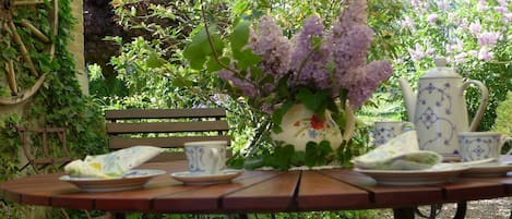Een tuintafel met een bloevas, een theepot en kopjes, omringd door groen en een bank.