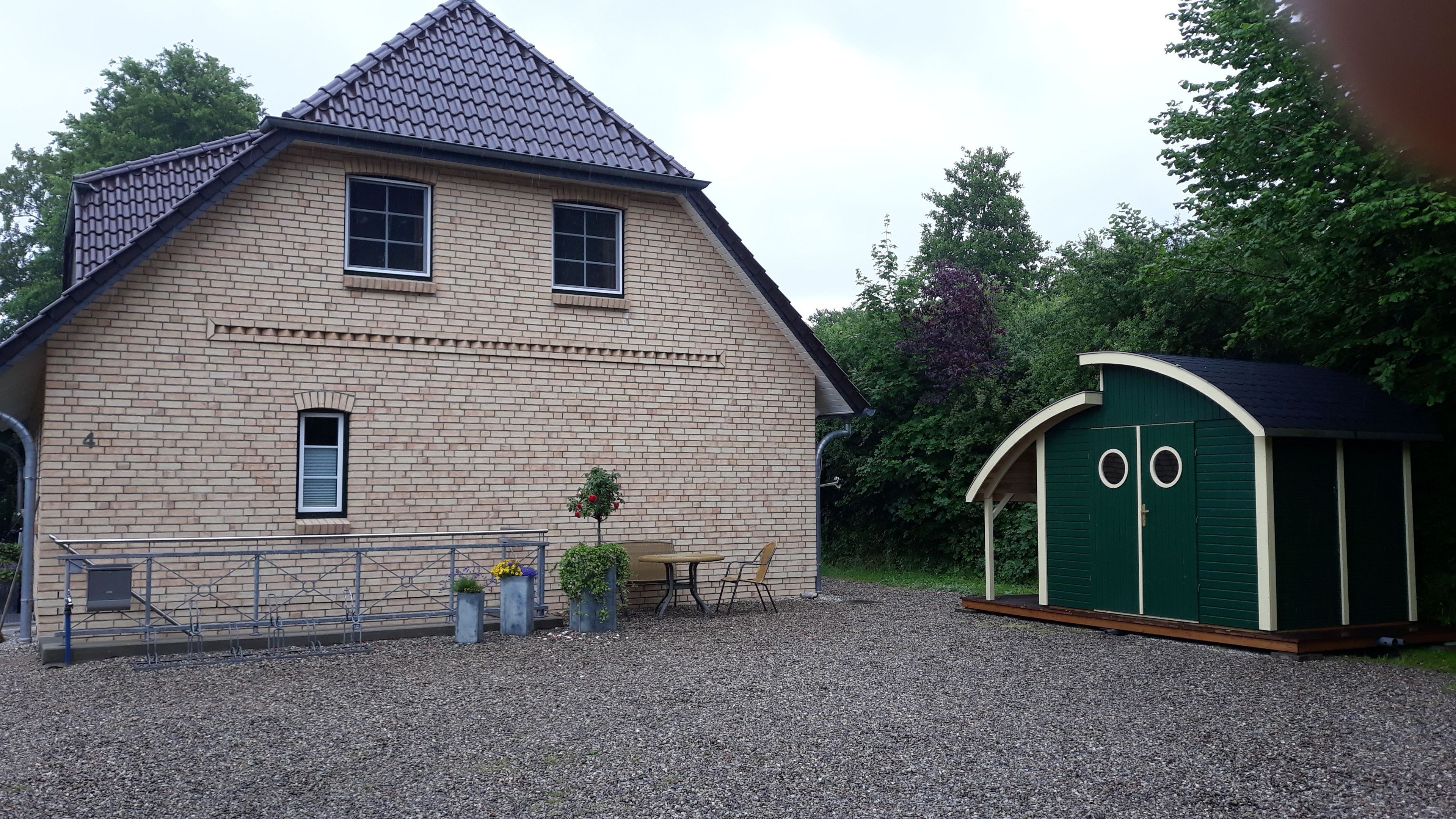 Ein zweistöckiges Haus mit Satteldach, einer kleinen Veranda mit Tisch und Stühlen und einem grünen Schuppen mit zwei Türen.