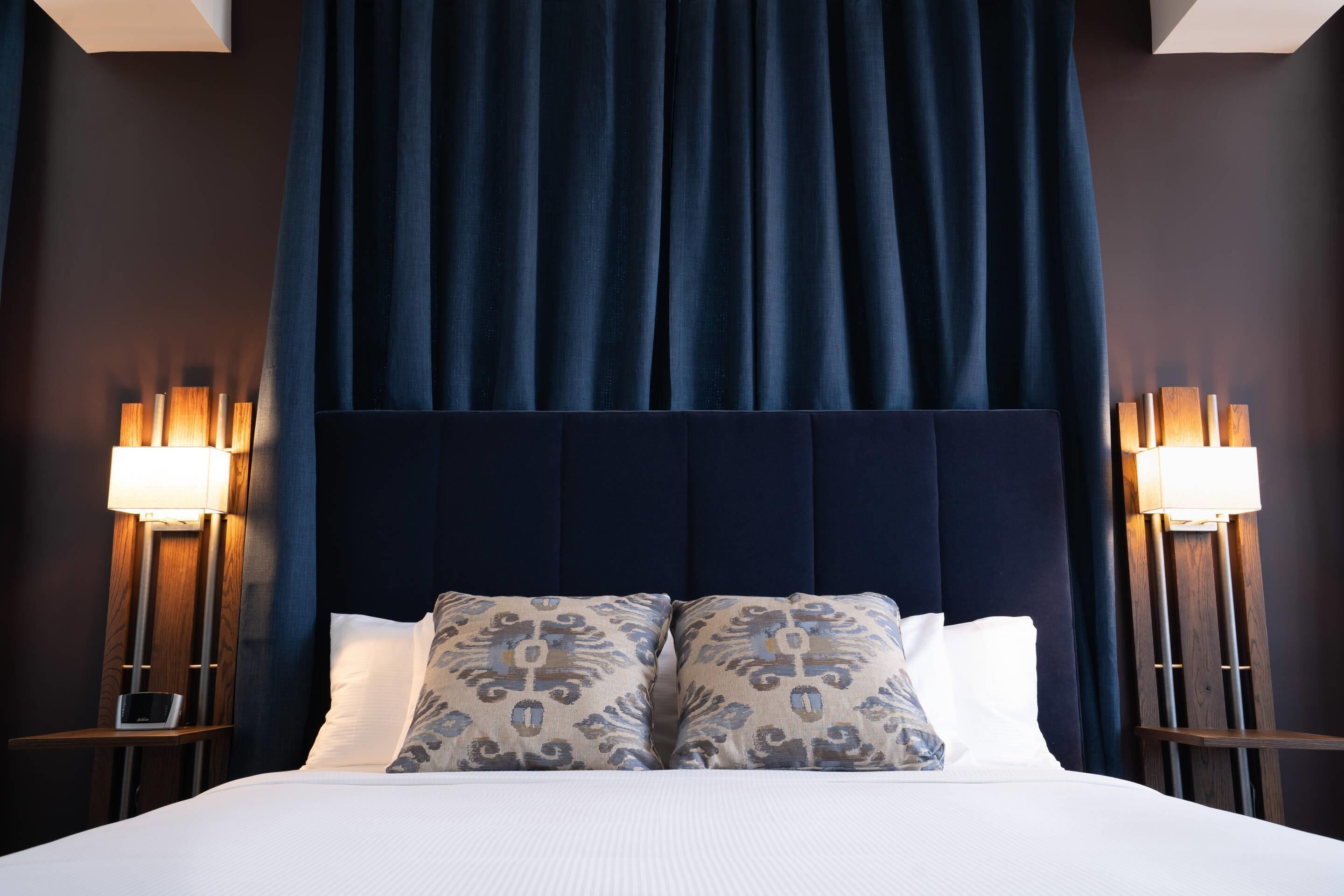 A hotel room with a large bed, dark headboard, two bedside lamps, and patterned pillows.