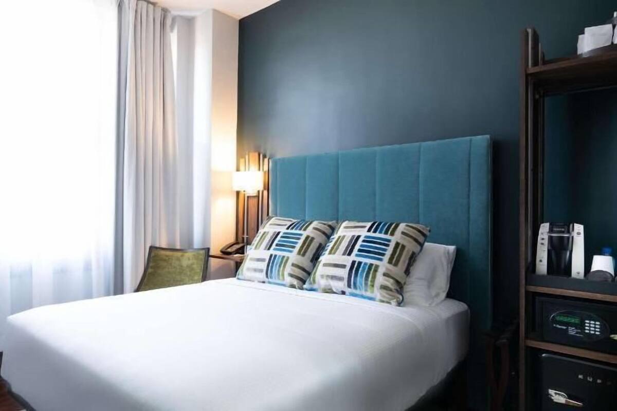 A hotel room with a bed, a nightstand, a lamp, a bookshelf, and a window with curtains.