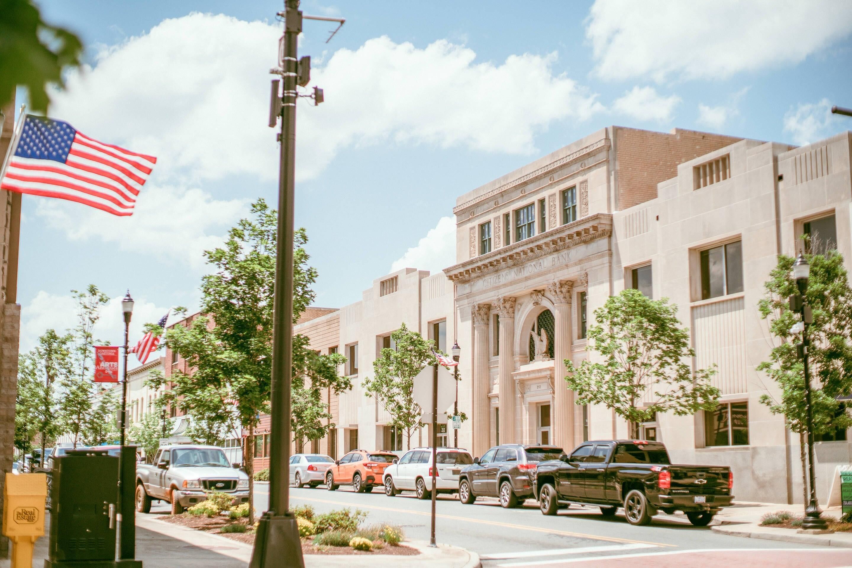 A street view with parked cars, a building with classical architecture, and an American flag.