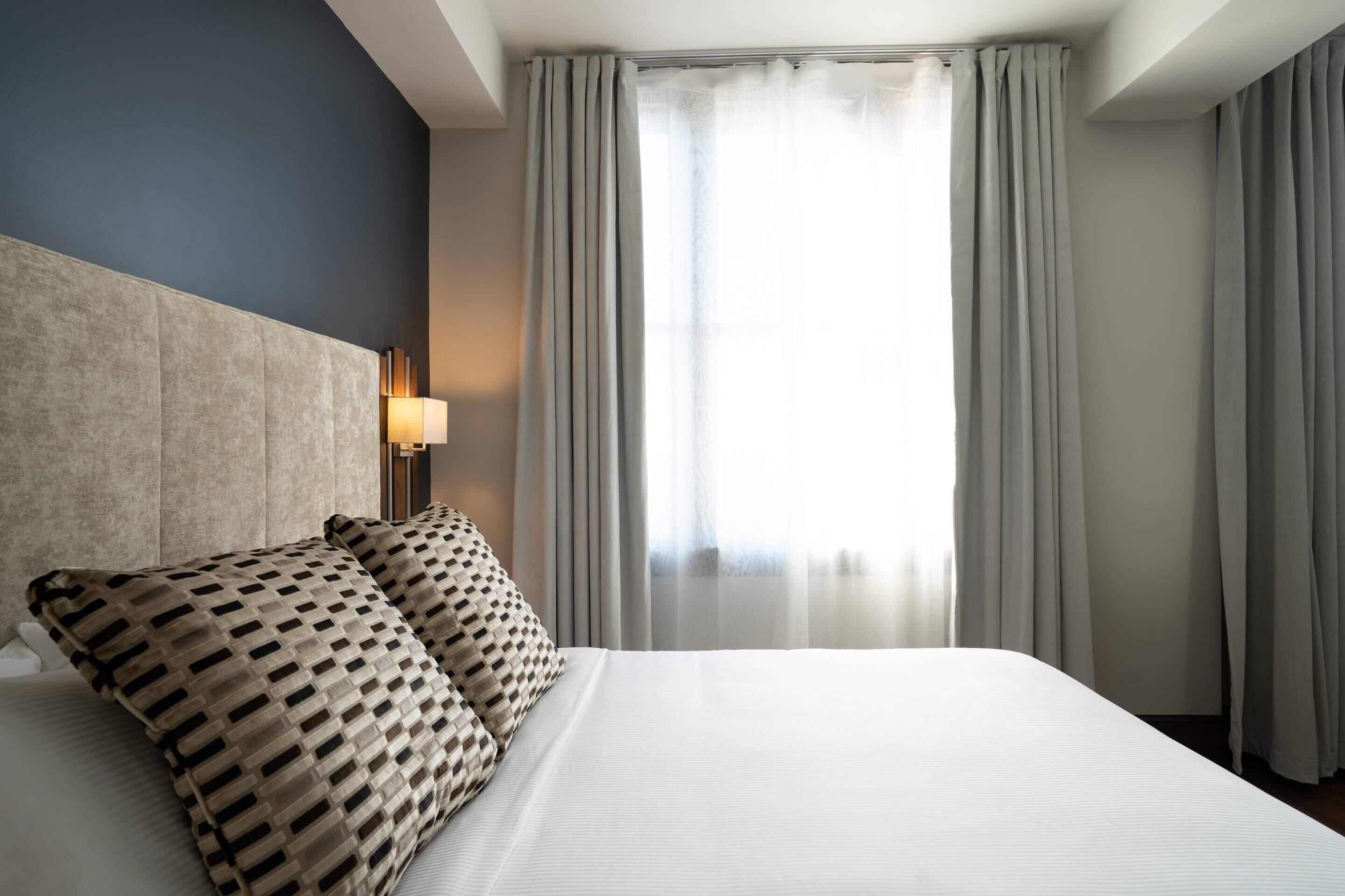 A hotel room with a large bed, a headboard, a bedside lamp, and a window with curtains.