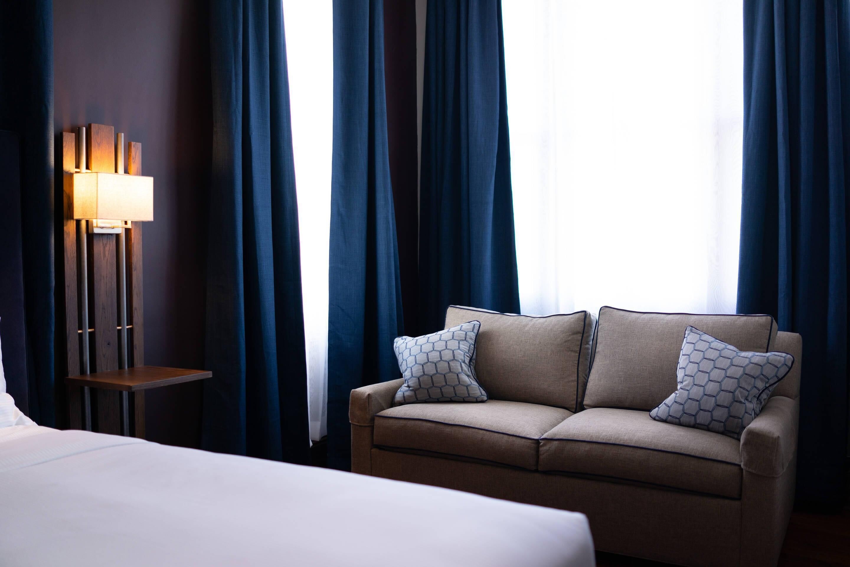 A hotel room with a bed, a sofa with patterned pillows, a standing lamp, and dark curtains.