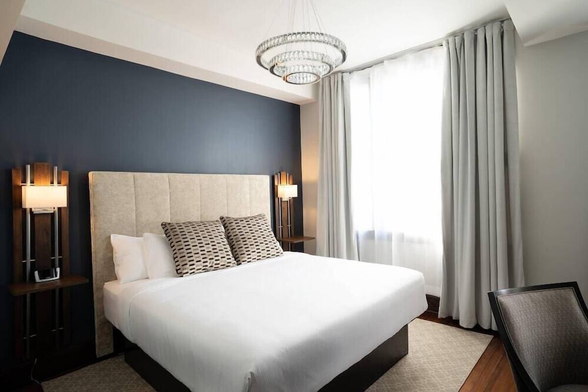 A modern hotel room with a large bed, two bedside lamps, a chandelier, and a view of the outdoors.