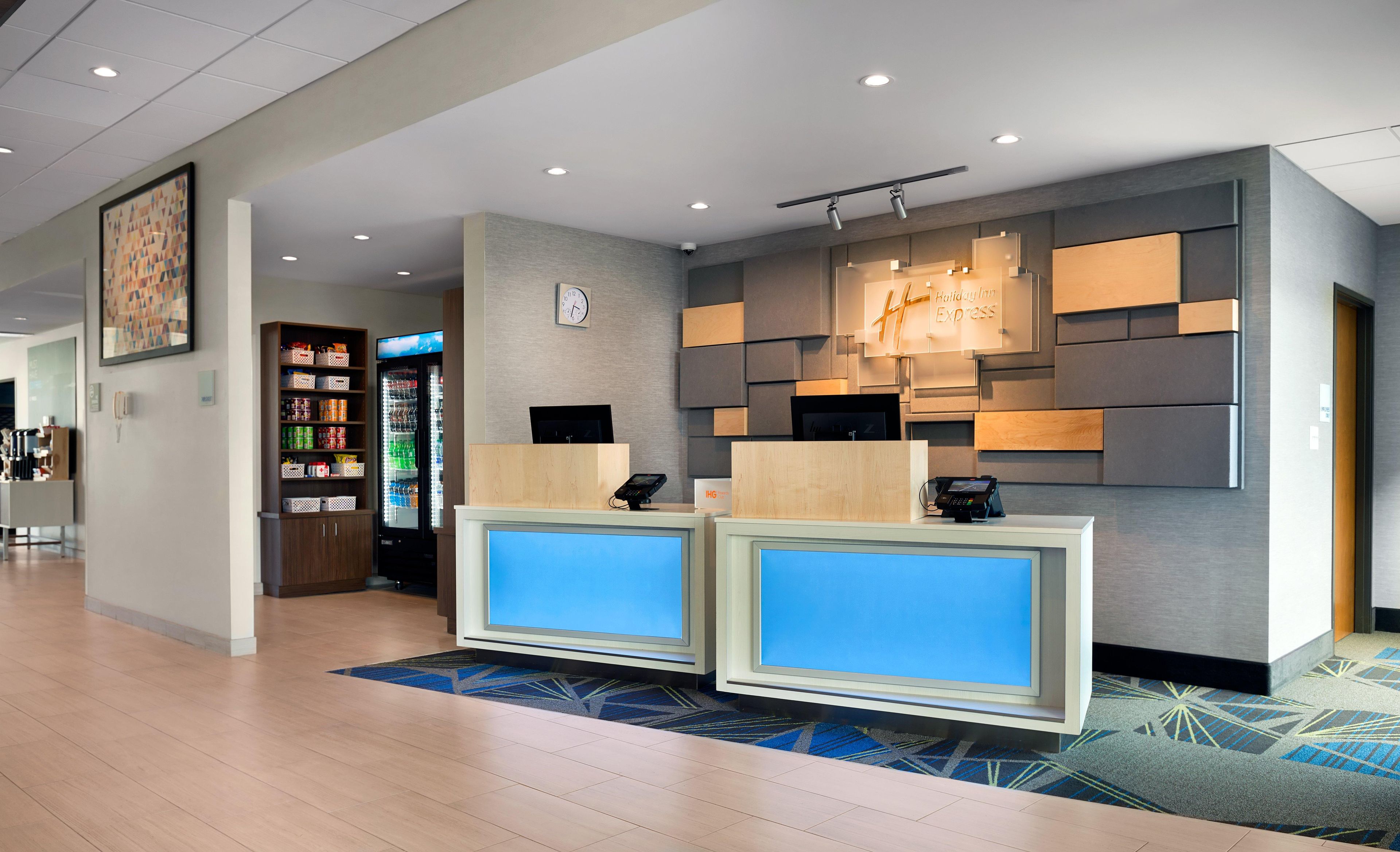 A hotel reception area with a modern design, including a digital display, a refrigerator stocked with beverages, and a clock on the wall.