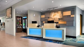 Ein Hotel-Rezeptionsbereich mit modernem Design, inklusive eines digitalen Displays, einem mit Getränken gefüllten Kühlschrank und einer Wanduhr.