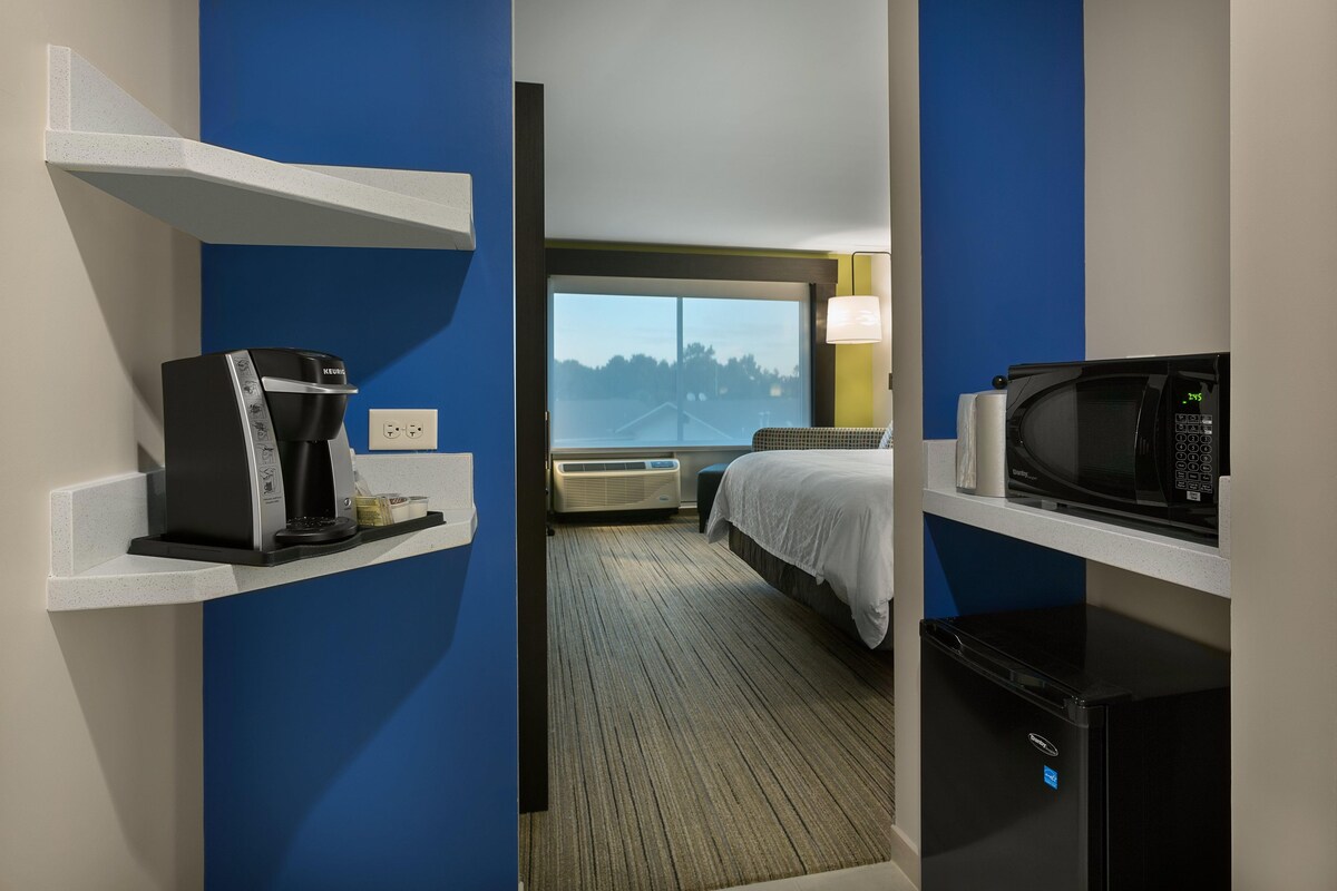 A hotel room with a bed, a coffee maker, a microwave, and a view of trees through a window.