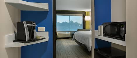 A hotel room with a bed, a coffee maker, a microwave, and a view of trees through a window.