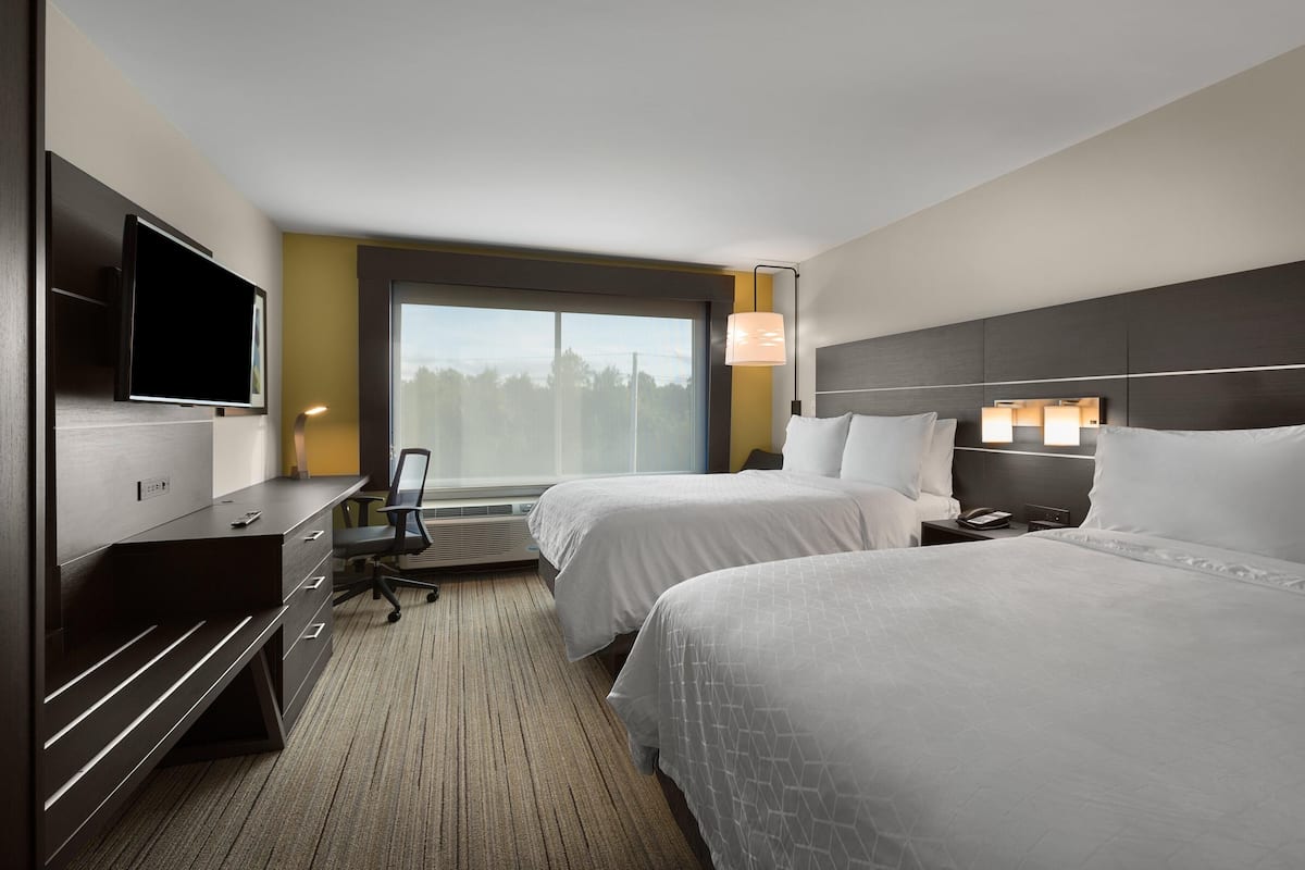 A hotel room with two beds, a desk with a chair, a flat-screen TV, a desk lamp, and a large window with a view of trees.