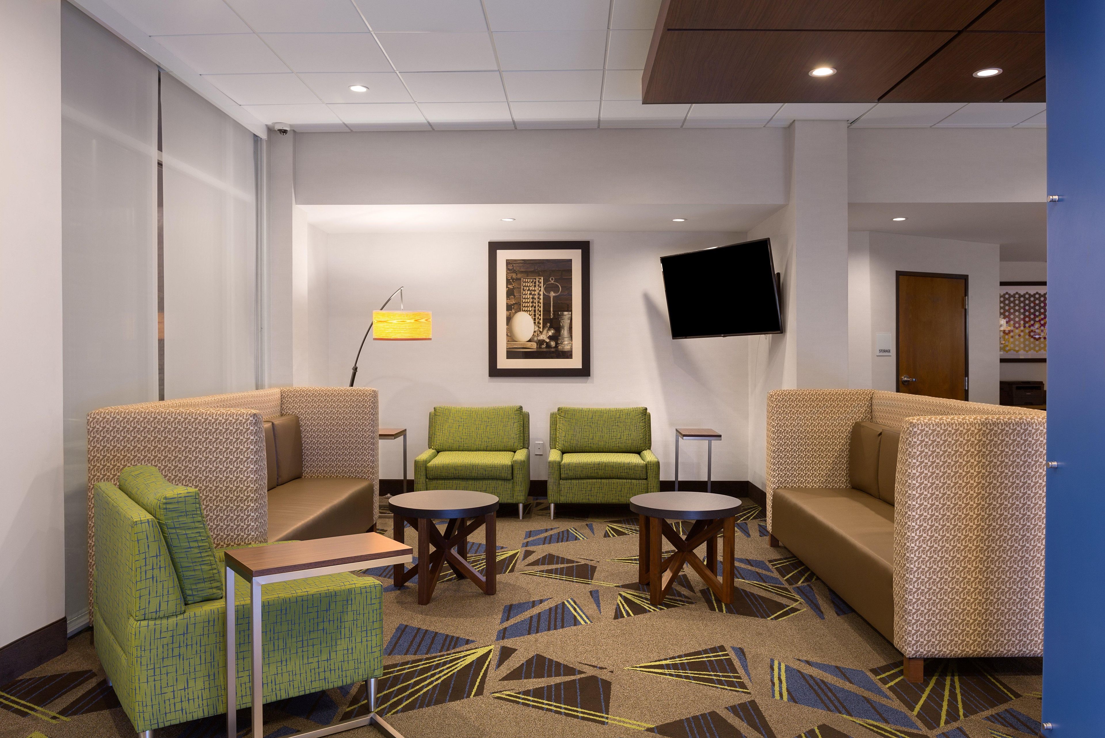 Un vestíbulo de hotel con un sofá, dos sillones, una mesita de centro, un televisor montado en la pared y una alfombra estampada.