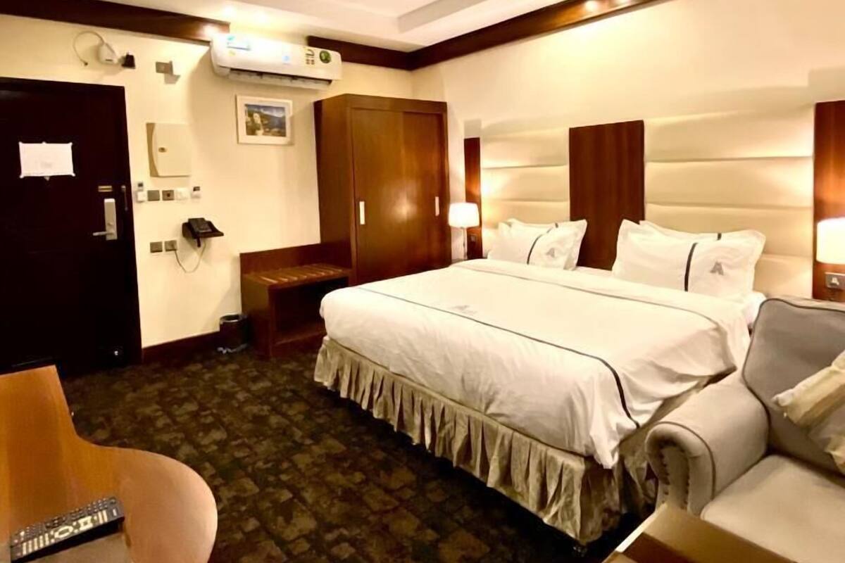 A hotel room with a large bed, a wooden wardrobe, a desk with a computer, and a small table with a glass object.