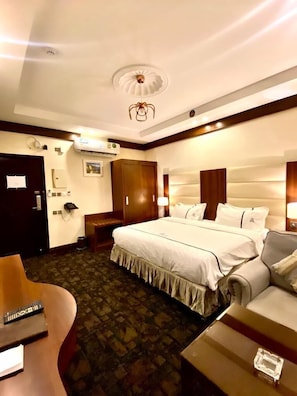 A hotel room with a large bed, a wooden wardrobe, a desk with a computer, and a small table with a glass object. - Aral Hotel (Tabuk)