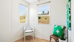 Ein Eckzimmer mit zwei Fenstern, ein Korbstuhl, ein Holzstuhl mit grünem Kissen und einer Topfpflanze.