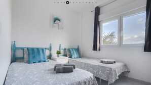 A bedroom with two beds, each with a patterned bedspread and matching pillows. There is a small table with a plant between the beds, and a window with curtains on the right side.