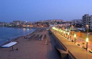 A coastal cityscape at dusk with a beach, promenade, and buildings in the background.
