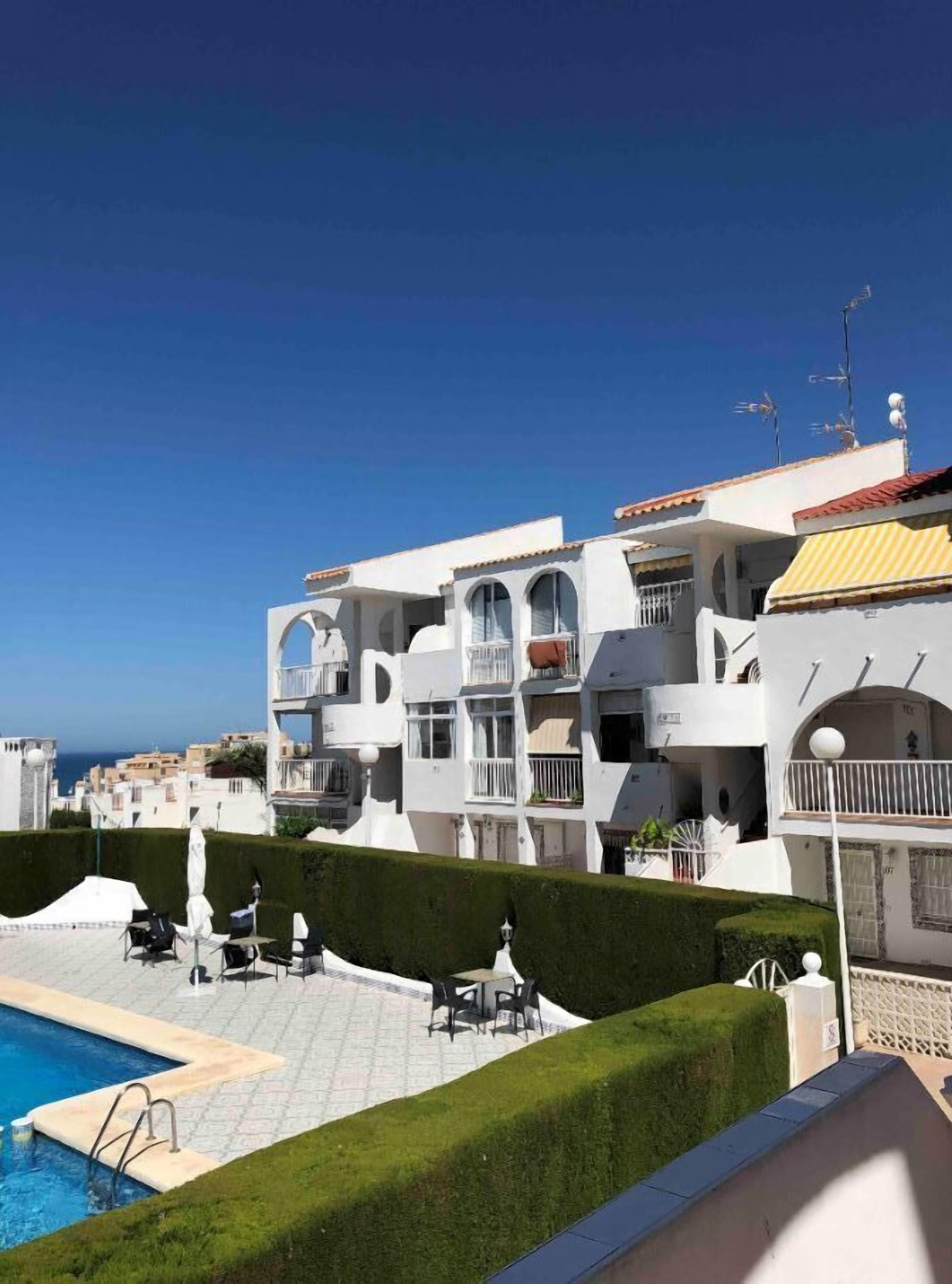 A pool area with a clear view of the sea, surrounded by white buildings with balconies and a well-maintained lawn.